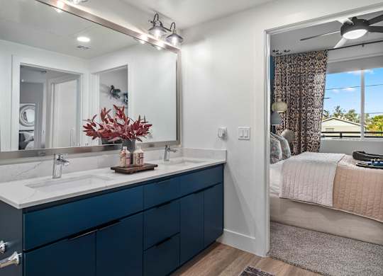 A bathroom with a large mirror, a sink, and a bedroom with a bed and a window.