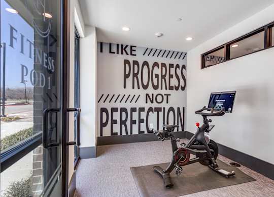 A gym with a wall that says "I like progress not perfection" and a stationary bike in front of it.