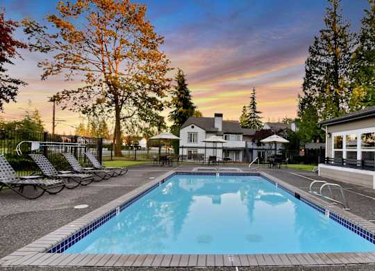 a swimming pool with chairs and a house with a sunset in the background