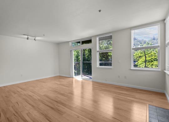 Spacious and bright living room with white walls, wood-style flooring, overhead lighting, and sliding glass door leading to private balcony at Collins Circle apartments in the Goose Hollow district in Portland, Oregon.