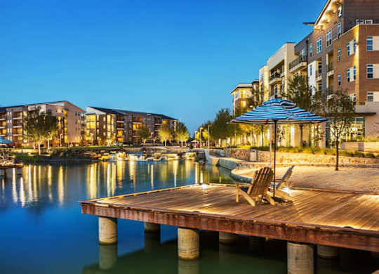 A wooden dock with chairs and an umbrella is in the foreground of a cityscape with buildings and boats.