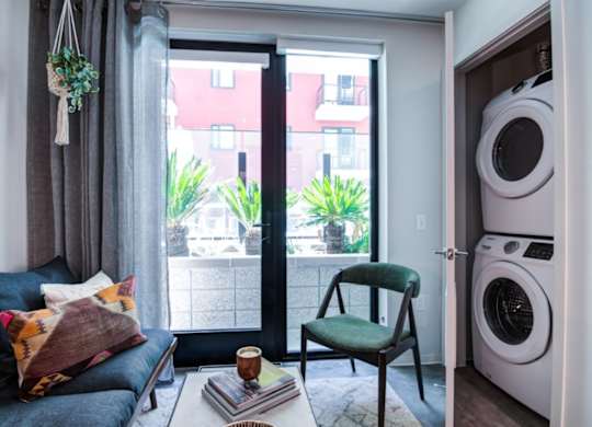 A living room with a grey couch, a green chair, a coffee table, and a view of the outside through a sliding glass door. and washer and dryer