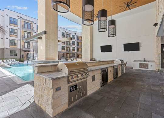 A stone BBQ grill is in the middle of a patio with a pool and apartment buildings in the background.