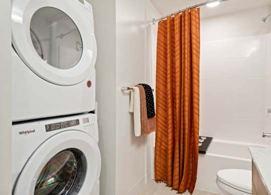 A white Whirlpool washing machine sits next to a bathroom with orange curtains.
