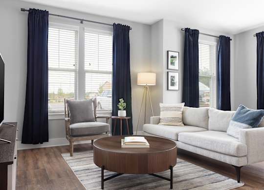 A living room with a grey sofa, a wooden coffee table, a grey armchair, a TV, and a window with blue curtains.