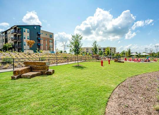 A sunny day at a park with a grassy area, trees, and apartment buildings in the background.