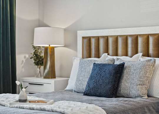 A bed with a white headboard and grey bedding with a lamp and vase on a nightstand.