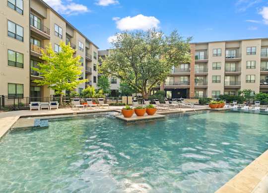 A swimming pool in front of apartment buildings with trees and lounge chairs.