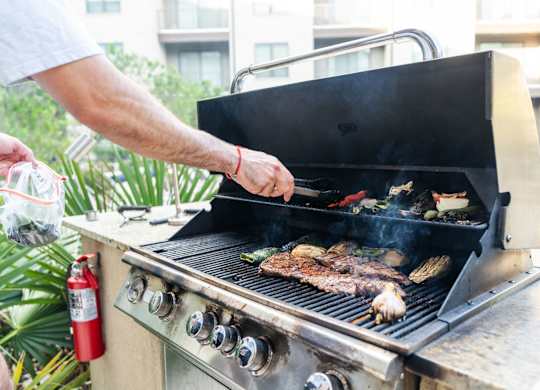 A man grilling meat on a barbecue with a red fire extinguisher nearby.