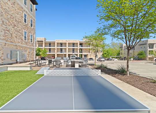 A shiny, metallic ping pong table is in the foreground of a sunny courtyard.