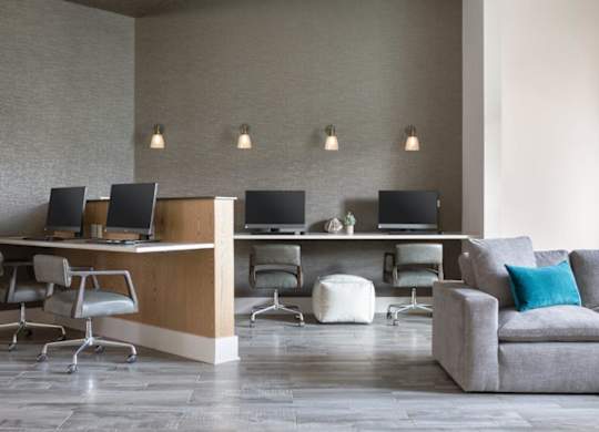 A modern office space with a grey couch, a wooden desk with two computers, and a white ottoman.