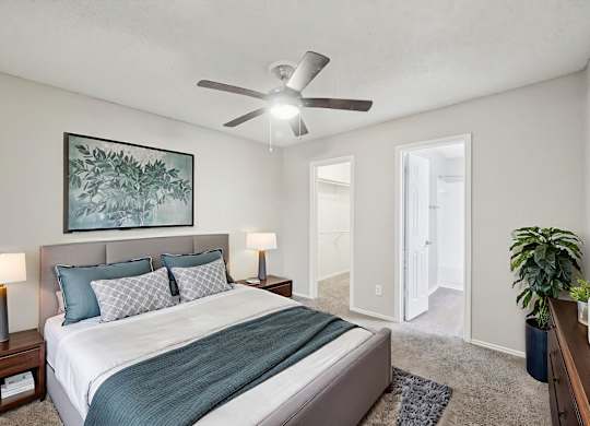 A bedroom with a bed, two lamps, a ceiling fan, and a painting on the wall.