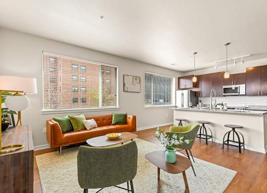 Modern living room with an orange couch, green accent chairs and oversized windows, complete with view into kitchen with barstool seating and pendant lighting at Sylvan Uptown, Denver, CO, 80203