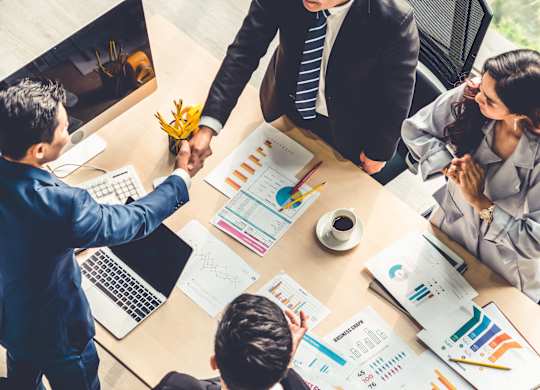 Three people shaking hands over a table with documents and graphs.