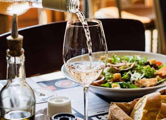 White wine being poured into a glass with a salad and bread on the table.