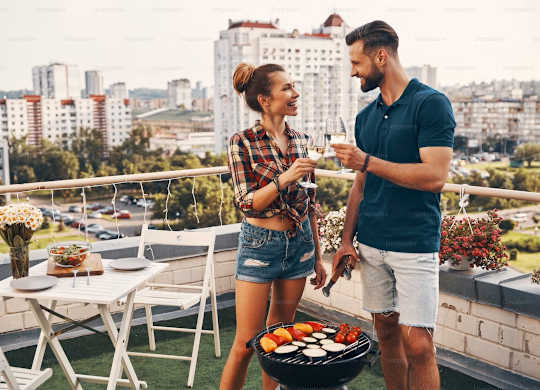 A man and woman are standing on a balcony with a barbecue grill in front of them.