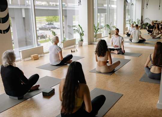 A group of people are sitting on yoga mats in a circle in a bright room.