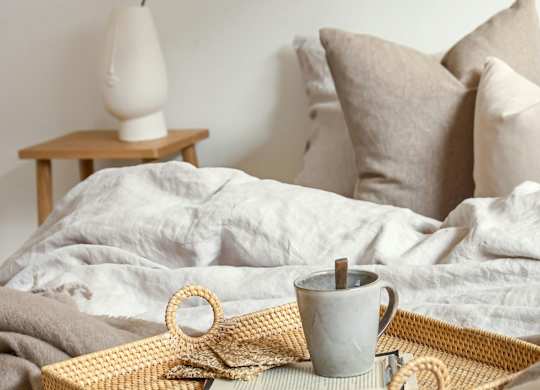 A grey mug with a wooden handle sits on a woven tray with a white lamp and pillows in the background.