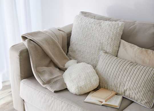 A beige couch with a white pillow, a white knitted hat, a book and a grey blanket.