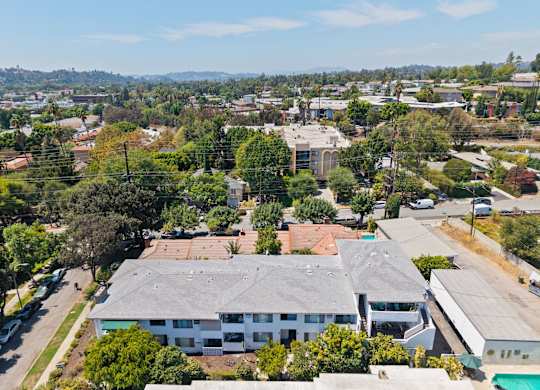 A large white building with a grey roof is surrounded by trees and other buildings.