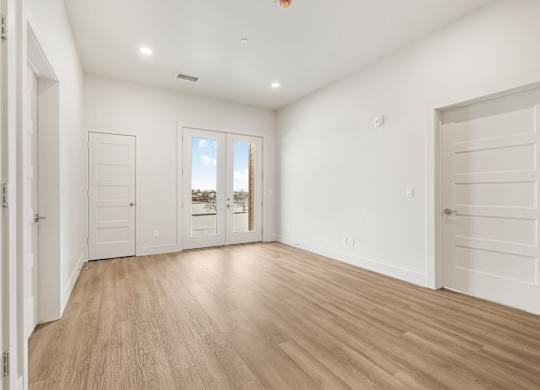 A room with white walls and wooden flooring, with a view of a building outside the window.
