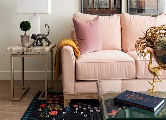 A living room with a pink couch, a small table with a plant and a cat figurine, and a rug with a colorful pattern.