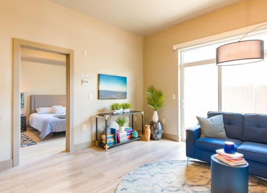 a living room with a blue couch and a lamp and a bedroom in the background at Sugar House Apartments By Urbana, Salt Lake City, UT, 84105
