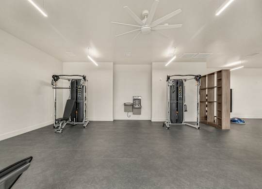 A gym with a grey floor and white walls has a fan and two pieces of gym equipment.