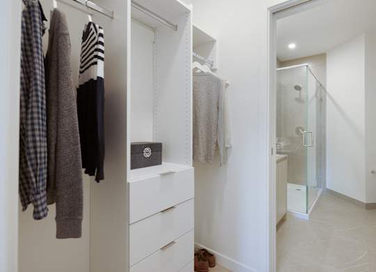 A white closet with clothes hanging and a bathroom in the background.