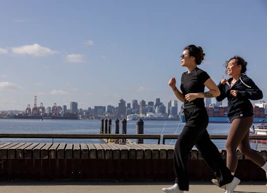 Two women jogging on a pier with a city skyline in the background.