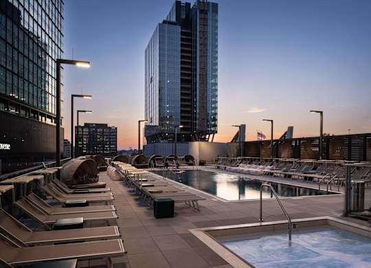 A rooftop pool area with lounge chairs and a hot tub in front of a city skyline at dusk.