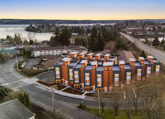 A large building with orange and blue exterior is surrounded by a road and trees. at The Narrows at Clare Apartments , Bremerton