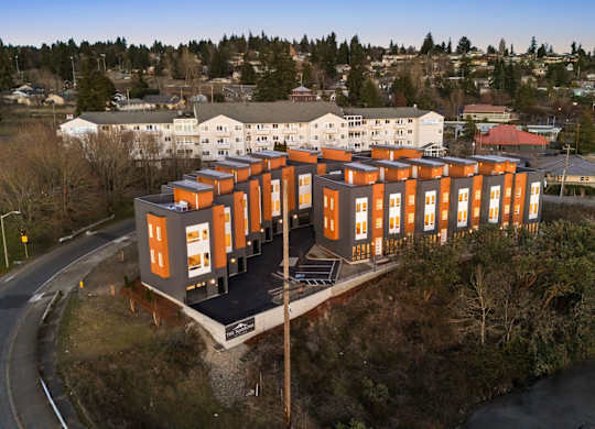 A modern building with orange and white exterior is situated on a small hill with a road and other buildings in the background. at The Narrows at Clare Apartments , Bremerton, 98310