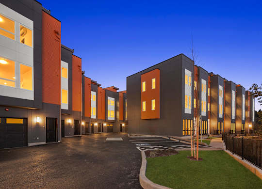 A modern building with orange and grey exterior is illuminated from within at The Narrows at Clare Apartments , Bremerton, WA, 98310