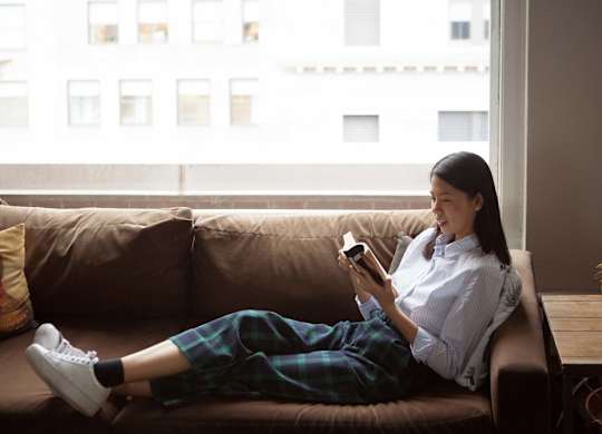 A woman is sitting on a couch with her legs crossed, wearing a white shirt and plaid pants, and holding a phone in her hands at B Flats Apartments, Bremerton, 98337