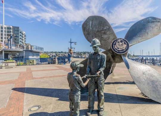 A statue of a man and a child are standing in front of a large propeller.
