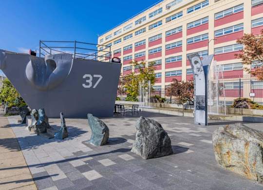 A large grey sculpture of a ship with the number 37 on it is in the foreground of a city square.