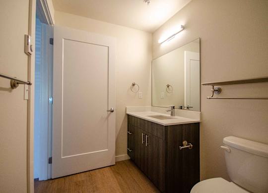 A bathroom with a white toilet, a sink, a mirror, and a white door at B Flats Apartments, Bremerton, WA, 98337