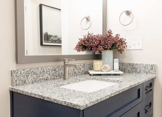 Primary bathroom vanity with a speckled granite countertop, blue cabinetry, and large mirror in a two-bedroom model apartment at The Crest at Oakwood in Oakwood, GA.