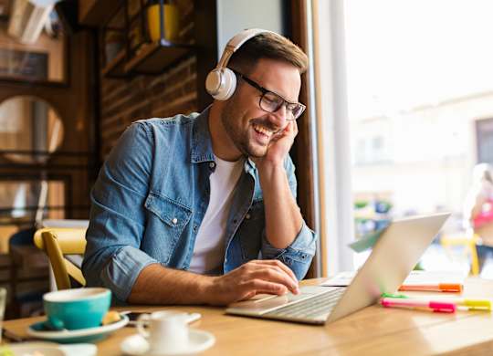 A man in a denim shirt is using a laptop and wearing headphones.