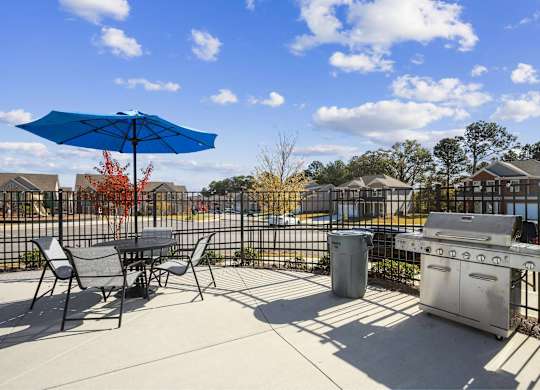 A blue umbrella is on a patio with a grill and chairs.