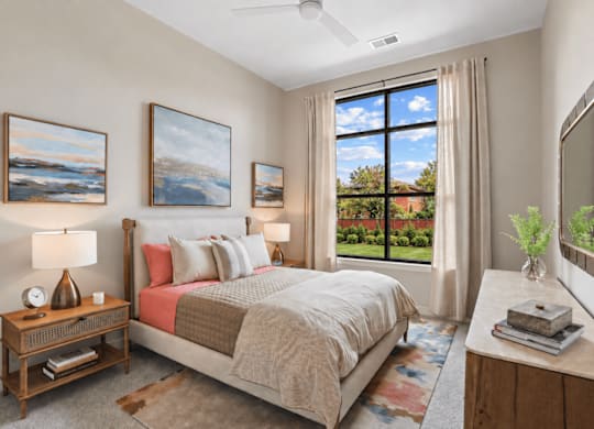 A bedroom with a large bed, a ceiling fan, and a view of the backyard through the window.