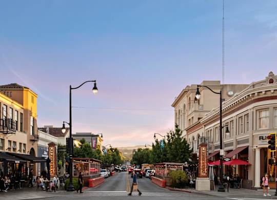 A street view of a city with buildings on either side and people sitting at tables outside the restaurants.