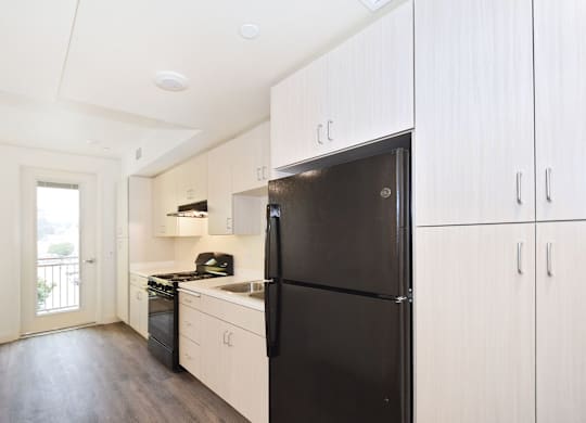 A black refrigerator stands in a kitchen with white cabinets and a wooden floor.