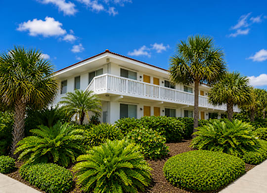 799 - A white building with a balcony is surrounded by green plants.