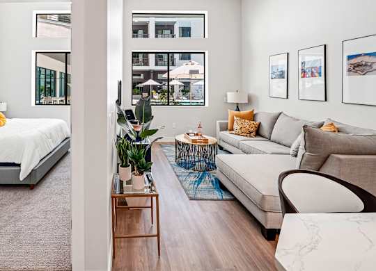 A modern living room with a grey sofa, a bed with white bedding, a wooden coffee table, and a large window with a view of the city.