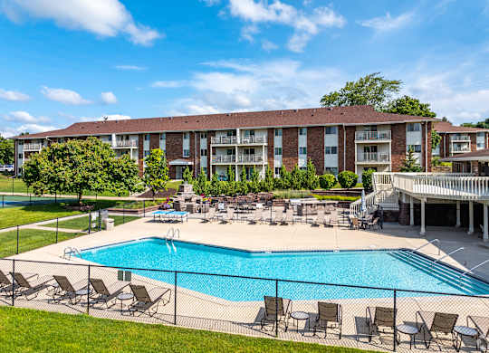 A large swimming pool surrounded by lawn chairs and umbrellas in front of a multi-story apartment building.