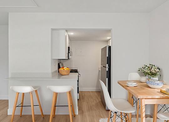 A kitchen with white walls and wooden floors, featuring a bar area with stools, a dining table with chairs, and a refrigerator.