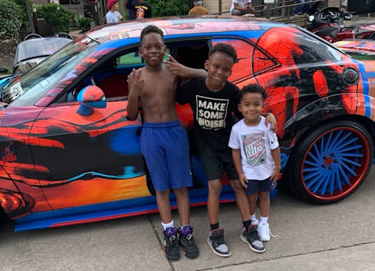 Two boys stand in front of a car with a black and orange design.