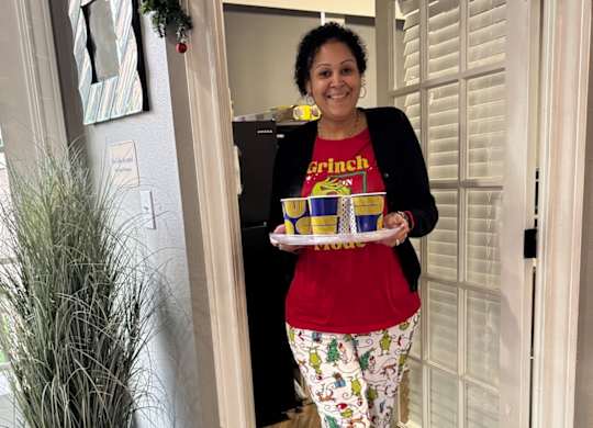 A woman in a red shirt and patterned pants holds a tray with a yellow box and a yellow cup.